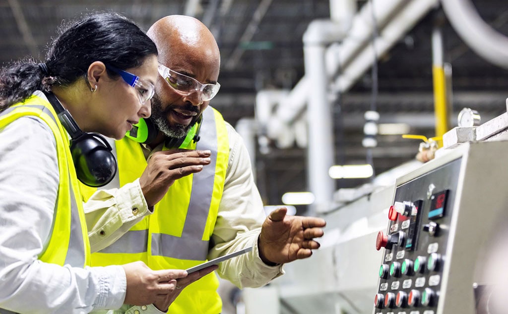 Two multi-ethnic workers working in a plastics factory, standing on the factory floor, looking at the control panel of one of the machines. The African-American man is pointing to the panel. His coworker, an Hispanic woman, is holding a digital tablet.